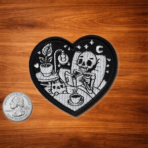 May include: Black heart-shaped patch with a white embroidered design of a skeleton reading in bed, with a plant, lamp, and coffee cup. A quarter coin is next to the patch. The patch has a black border.