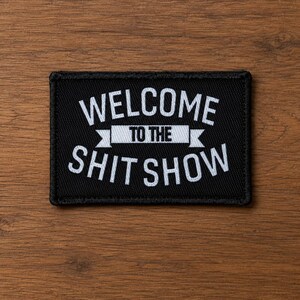 Può includere: Una toppa rettangolare nera ricamata con un bordo bianco. Il testo WELCOME TO THE SHIT SHOW è ricamato in lettere maiuscole bianche, con le parole TO THE centrate su un grafico a forma di striscione bianco tra le due righe di testo.