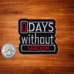 以下が含まれることがあります： 白と赤で「0 DAYS without SARCASM」の文字が刺繍された黒いパッチ。サイズの比較のために、パッチの隣に米国の25セント硬貨があります。パッチは木製の表面にあります。