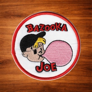 Puede incluir: Un parche bordado circular que presenta al personaje Bazooka Joe con una gorra negra y soplando una gran burbuja rosa. El texto BAZOOKA está bordado en rojo sobre el personaje y la palabra JOE está bordada en rojo debajo del personaje.