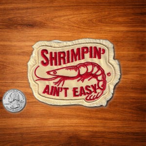 May include: Embroidered patch with the words "SHRIMPING AIN'T EASY" in red, along with a red shrimp illustration. The patch has a cream-colored background and a decorative border. A quarter is next to the patch.