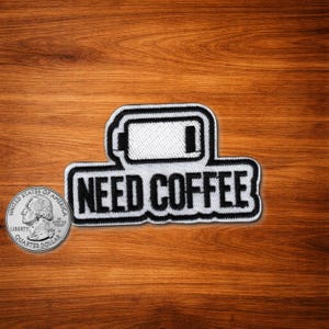 May include: A white patch with black border featuring a battery icon and the words "NEED COFFEE" in bold, black letters. The patch is placed on a wooden surface next to a quarter.