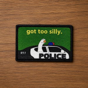 Peut inclure: Un écusson rectangulaire brodé sur fond vert représentant une oie blanche au bec orange sortant d'une voiture de police noire et blanche. Le texte indique "got too silly.", "911" et "POLICE" sur la portière du véhicule.