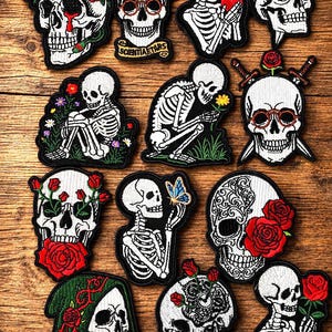 May include: A collection of thirteen embroidered patches featuring white skulls and skeletons. The designs include skulls adorned with red roses, skeletons holding hearts, flowers, or butterflies, and skulls wearing glasses or hoods, all with black stitched borders.