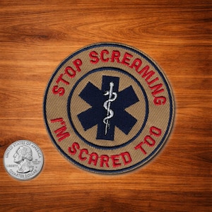 May include: A round patch with the text "STOP SCREAMING I'M SCARED TOO" in red lettering. The patch features a blue star of life symbol with a white caduceus in the center. The patch is tan with a blue border.
