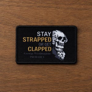 Puede incluir: Un parche rectangular negro con un gráfico en blanco y dorado de una calavera con el peinado de George Washington. El texto dice: STAY STRAPPED OR GET CLAPPED -George Washington- PROBABLY. El parche tiene un borde negro cosido.