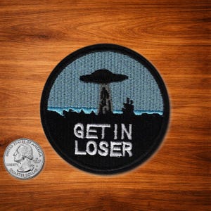 May include: Round black patch with the text "GET IN LOSER" in white. The patch features a blue sky background with a UFO and a landscape silhouette. A quarter is next to the patch.
