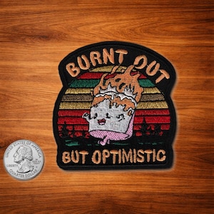 May include: Embroidered patch with the words "BURNT OUT BUT OPTIMISTIC" above and below a cartoon cupcake with flames. The patch has a black border and a colorful sunset design.