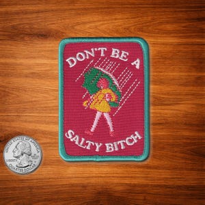 Puede incluir: Parche bordado con el texto "DON'T BE A SALTY BITCH" y una ilustración de una persona bajo un paraguas bajo la lluvia. El parche tiene un fondo rojo con un borde turquesa.
