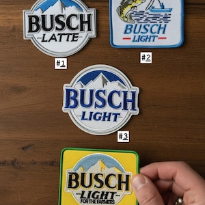 May include: Four embroidered fabric patches featuring Busch branding. Patch 1 is circular with a mountain graphic and text BUSCH LATTE. Patch 2 is rectangular with a fish and boat graphic and text BUSCH LIGHT. Patch 3 is circular with mountains and text BUSCH LIGHT. Patch 4 is yellow and rectangular with mountains and text BUSCH LIGHT FOR THE FARMERS.