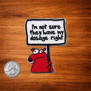 May include: Embroidered patch with a red cartoon character holding a sign. The sign reads "I'm not sure they have my dosage right" in black text on a white background. The patch is bordered with black stitching. A quarter is in the lower left corner.