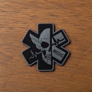 May include: A six-pointed star-shaped patch in dark gray and black. The design features a split graphic with a skull on the left side and a heartbeat line with the word MEDIC on the right side. The background of the patch has a textured honeycomb pattern.