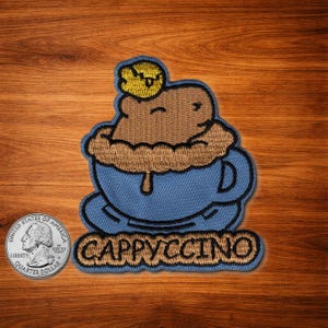 Puede incluir: Parche bordado con un diseño de capuchino. El parche muestra una taza de café azul con una bebida de café marrón y una cobertura amarilla. La palabra "CAPPUCCINO" está bordada en marrón en la parte inferior.