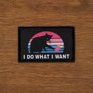 May include: A rectangular black patch featuring a graphic of a black cat reaching towards a pink cup against a sunset background of horizontal pink, purple, and blue stripes. The text I DO WHAT I WANT is printed in white capital letters below.
