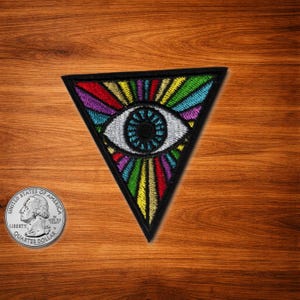 Könnte beinhalten: Ein farbenfroher, gestickter Aufnäher in Dreiecksform mit einem Augendesign mit Regenbogenstrahlen. Das Auge hat eine schwarze Pupille und eine weiße Iris. Der Aufnäher ist schwarz umrandet und vor einem Holzgrund platziert.