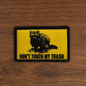 May include: A rectangular embroidered patch with a yellow background and a black border. The patch features a black illustration of a raccoon in the center and the text DON'T TOUCH MY TRASH printed in black capital letters below the raccoon.