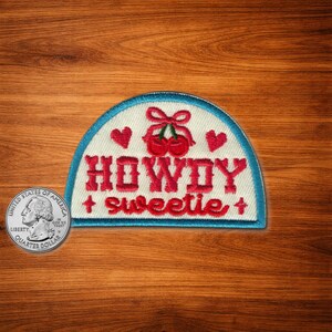 Puede incluir: Parche bordado con fondo blanco, borde azul y las palabras "HOWDY sweetie" en rojo. El parche presenta corazones rojos y cerezas con un lazo.
