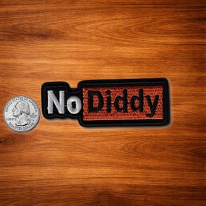 May include: A black embroidered patch with the words "No Diddy" in white and black. The patch has a rectangular shape with a red background and a black border. A U.S. quarter is next to the patch for size comparison.