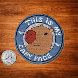 May include: A round, blue embroidered patch with the text "THIS IS MY CAPY FACE" in white. The patch features a cartoon capybara face with a brown body, red nose, and black eye. A quarter is next to the patch.