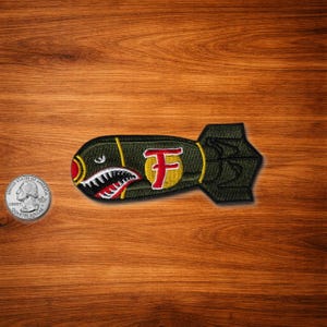 May include: Embroidered patch in the shape of a bomb, with a shark mouth design in red, white, and black. The patch has a green body with a yellow and red letter "F" and yellow accents. A quarter coin is next to the patch.