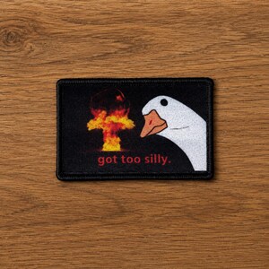 Puede incluir: Un parche rectangular bordado con fondo negro que muestra una cabeza de ganso blanca a la derecha, una nube de hongo naranja y amarilla a la izquierda y el texto rojo "got too silly." impreso en la parte inferior central.
