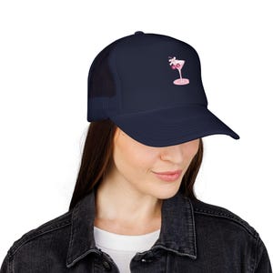 Puede incluir: Una gorra de camionero azul marino con parte trasera de malla y panel frontal sólido. La parte delantera de la gorra muestra un pequeño gráfico centrado de una copa de cóctel rosa claro con un lazo blanco y dos cerezas rojas oscuras.