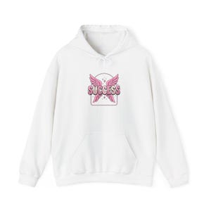 Pode incluir: Moletom branco com bolso frontal e capuz com cordão. A frente apresenta um gráfico rosa e branco de asas e a palavra "SUCCESS". O moletom é feito de um material macio e confortável.