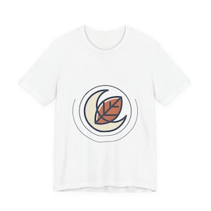 May include: White t-shirt with a circular design. The design features a cream-coloured crescent moon, a rust-coloured leaf, and a navy blue outline. The design is centered on the front of the shirt, offering a minimalist aesthetic.