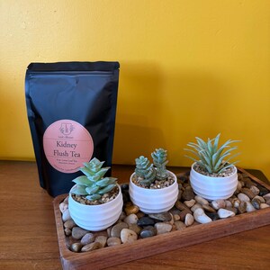 May include: A black pouch labeled Kidney Flush Tea with a pink circular sticker, positioned next to a rectangular wooden tray filled with smooth river stones and three small white ribbed pots containing green artificial succulent plants.