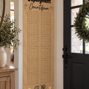 May include: A vertical tan fabric wall hanging featuring the text Daddy's Deployment Countdown in black. It includes two columns of grid squares for tracking days, decorated with small envelope icons, hanging stars, and illustrations of dog tags and boots.
