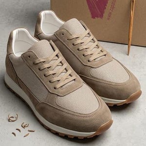 Puede incluir: Zapatillas beige de ante y malla con suelas blancas y detalles marrones. Una caja de cartón con un logotipo "V" burdeos y el sitio web "zapatosvias.com" en el fondo. Calzado sobre una superficie gris.