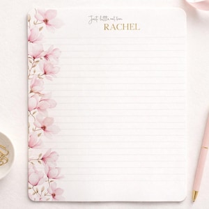 Puede incluir: Un bloc de notas rayado con un diseño floral con flores de magnolia rosas. El texto "Just little notes from RACHEL" está en dorado. También hay un bolígrafo rosa y un cuenco pequeño con clips dorados.