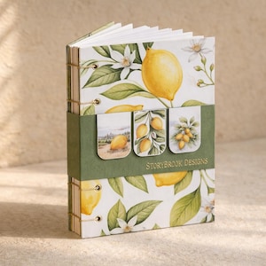 Könnte beinhalten: Ein dekoratives Notizbuch mit Zitronen-Motiv. Das Cover und die Seiten zeigen Illustrationen von Zitronen, Blättern und Blüten. Ein grünes Band umschließt das Notizbuch, auf dem der Text "Storybrook Designs" aufgedruckt ist. Das Notizbuch ist mit Bindfaden gebunden.