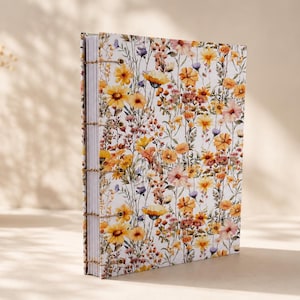 May include: A rectangular hardback journal featuring a white cover with a dense pattern of yellow, orange, and muted purple wildflowers. The book is bound with visible exposed stitching along the spine, showcasing white pages stacked neatly inside.