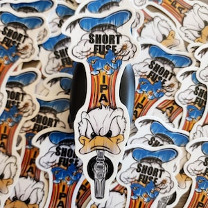 Könnte beinhalten: Eine Ansammlung weißer Aufkleber mit einer Comic-Ente mit wütendem Gesichtsausdruck. Die Aufkleber tragen die Wörter "SHORT FUSE" und "IPA" in einem stilisierten Design mit blauen und orangefarbenen Akzenten. Die Ente hält einen Zapfhahn.