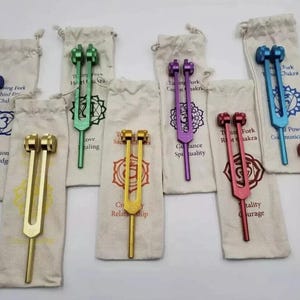 May include: A selection of vibrantly coloured tuning forks, including blue, green, purple, gold, and red, each with a matching beige drawstring bag. The bags have text and chakra symbols. These forks are used for sound therapy and meditation.