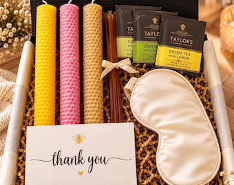 May include: A gift box containing three cylindrical honeycomb-textured beeswax candles in yellow, pink, and tan, a bundle of honey sticks, two Taylors of Harrogate green tea bags, a cream-colored sleep mask, and a thank you card with a bee icon.