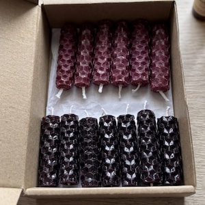 May include: A box containing two rows of decorative candles. The top row has burgundy candles, the bottom row has dark purple candles. The candles have a honeycomb pattern. Two essential oil bottles and crystals are in the background.