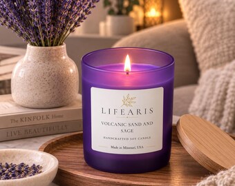May include: A purple frosted glass candle with a lit wick and a white label reading "LIFEARIS VOLCANIC SAND AND SAGE HANDCRAFTED SOY CANDLE Made in Missouri, USA". The candle sits on a round wooden tray next to a small bowl of dried lavender.