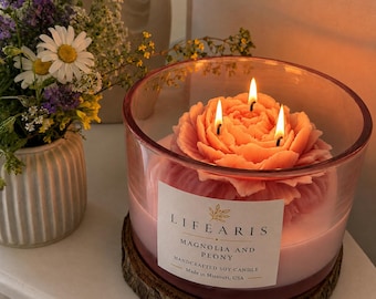May include: A three-wick soy candle shaped like a pink peony flower inside a clear glass jar. The white label reads "LIFEARIS MAGNOLIA AND PEONY HANDCRAFTED SOY CANDLE Made in Missouri, USA". The candle sits on a circular wooden base.