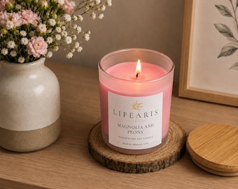May include: A pink soy candle in a clear glass jar with a white label reading "LIFEARIS MAGNOLIA AND PEONY HANDCRAFTED SOY CANDLE Made in Missouri, USA". The candle has a lit wick and sits on a circular wooden coaster next to a wooden lid.