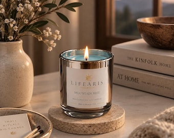 May include: A lit soy candle in a reflective silver glass container with a white label reading "LIFEARIS MOUNTAIN MIST HANDCRAFTED SOY CANDLE Made in Missouri, USA" sitting on a round speckled stone coaster with a visible flame and blue wax.