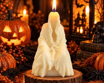 May include: A cream-colored wax candle shaped like a draped figure or ghost, featuring a lit wick at the top. The candle is positioned on a circular wooden slice base, displaying detailed folds and textures in the wax that mimic flowing fabric.