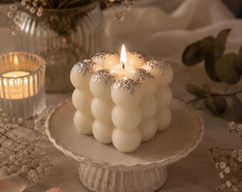 May include: A square bubble candle made of off-white wax, featuring a lit wick in the center and silver leaf accents applied to the top spheres. The candle is positioned on a small, round, textured ceramic pedestal stand with a scalloped edge.