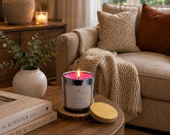 May include: A lit candle in a reflective silver glass jar with a white label reading "LIFEARIS JAPANESE CHERRY BLOSSOM". The candle sits on a small wooden coaster next to a round bamboo lid, positioned on a wooden table with books.