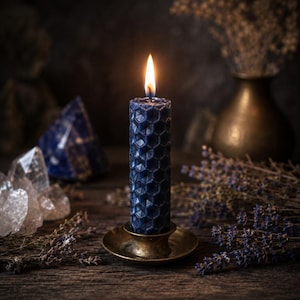 May include: A dark blue cylindrical candle with a raised hexagonal honeycomb texture, featuring a lit wick with a bright flame. The candle is set in a small, circular, metallic brass-toned holder, creating a warm glow from the flame.