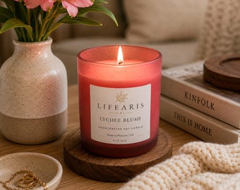 May include: A lit red soy candle in a glass jar with a white label reading "LIFEARIS LYCHEE BLUSH HANDCRAFTED SOY CANDLE 8 OZ, 227g". The candle sits on a round wooden coaster next to a speckled ceramic vase with pink flowers and a small jewelry dish.