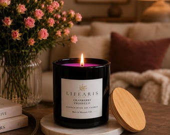 May include: A black glass candle jar with a white label reading LIFEARIS CRANBERRY PROSECCO HANDCRAFTED SOY CANDLE Made in Missouri, USA. The candle has a lit wick with a bright flame and sits on a white marble coaster next to a round wooden lid.