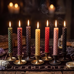 May include: Seven lit, colorful, honeycomb-patterned candles in brass holders. The candles are in shades of green, red, purple, yellow, red, pink, and black. Smoke rises from the candles, and the background is dark.