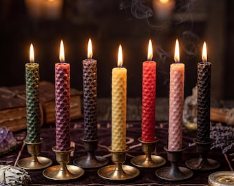 May include: Seven lit, colorful, honeycomb-patterned candles in brass holders. The candles are in shades of green, red, purple, yellow, red, pink, and black. Smoke rises from the candles, and the background is dark.